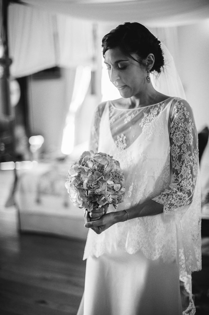 Black and white portrait of bride in lace wedding dress holding floral bouquet indoors.