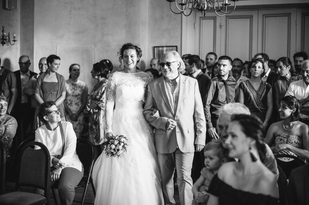 Black and white photo of a bride in a lace gown being escorted down the aisle by a man.