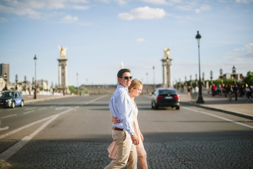 Couple walking across a Parisian street with ornate bridge and golden statues in background.