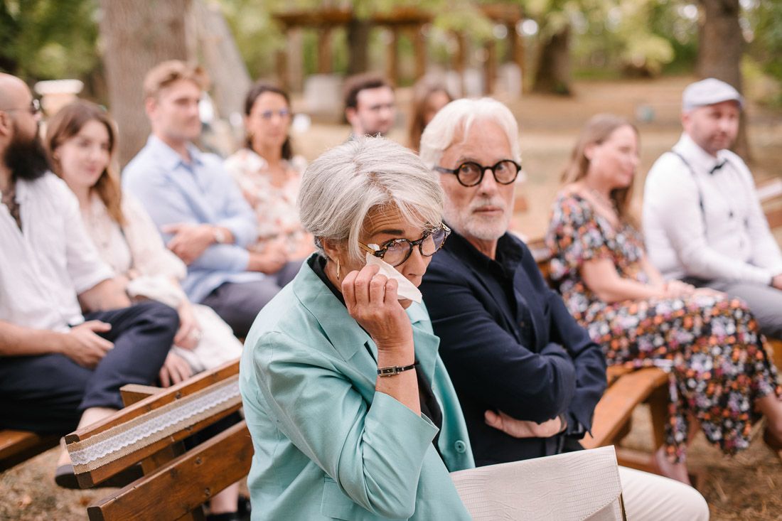 Emotional wedding guests seated outdoors, older woman wiping tears while man in glasses sits beside her.