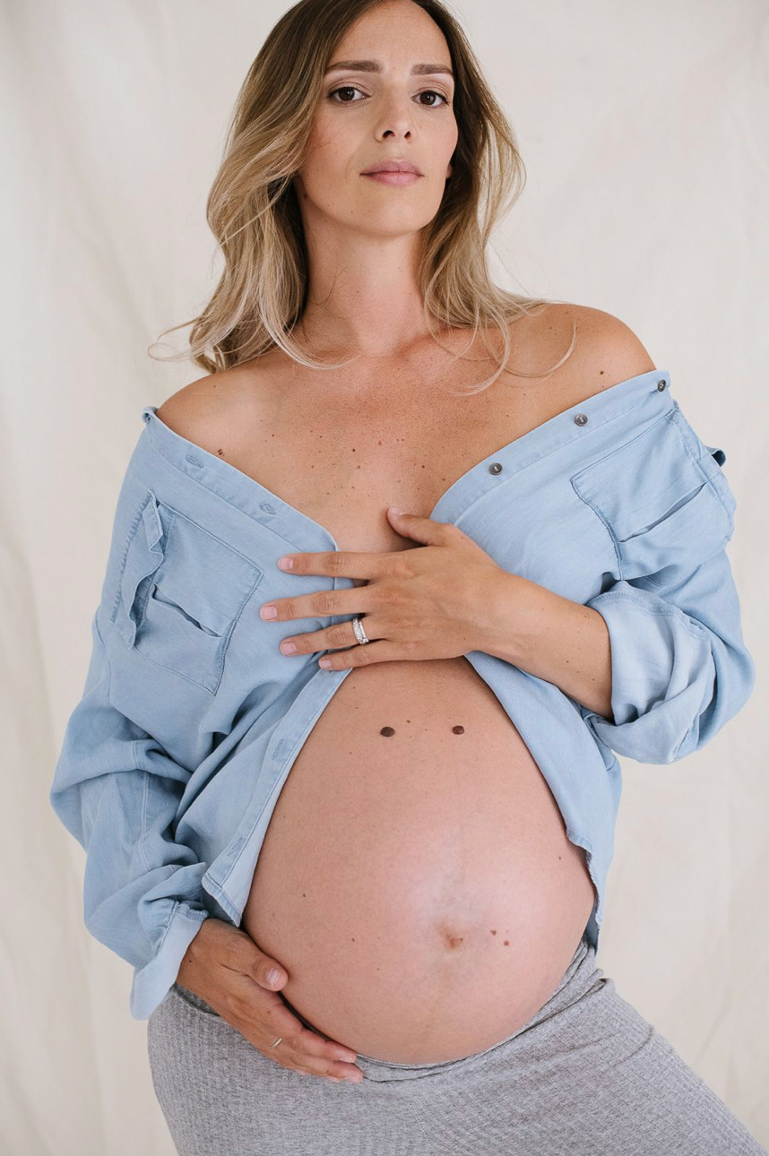 Pregnant woman in open blue shirt cradling her bare baby bump during a maternity photo session.
