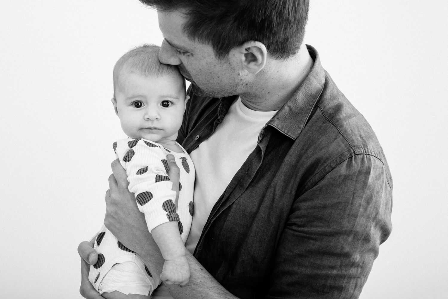 Father kissing newborn baby on forehead in black and white portrait.