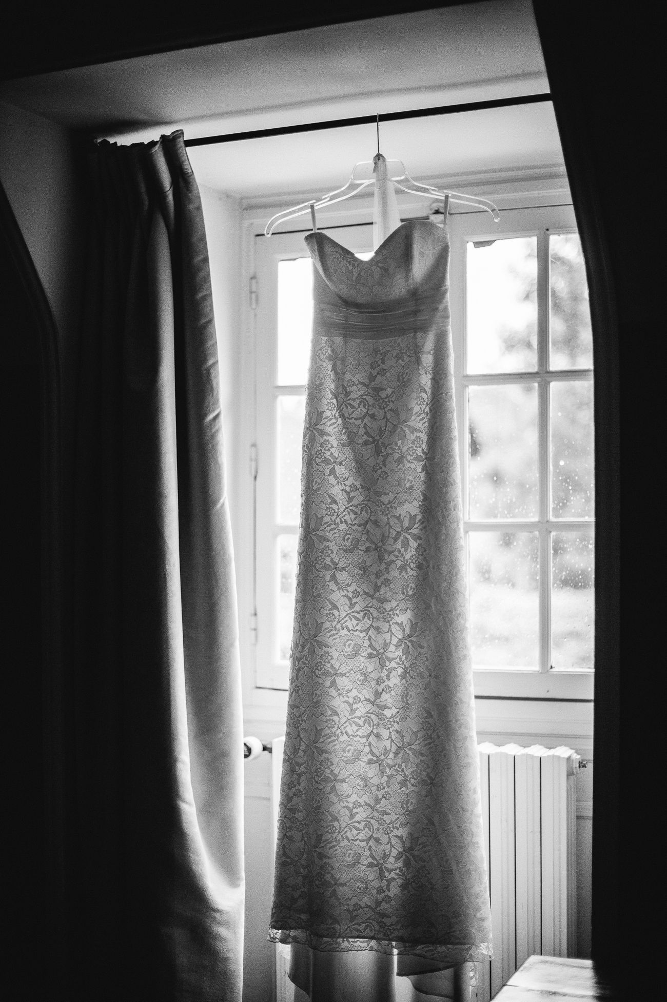 Elegant strapless lace wedding dress hanging on a hanger in front of a window in black and white.