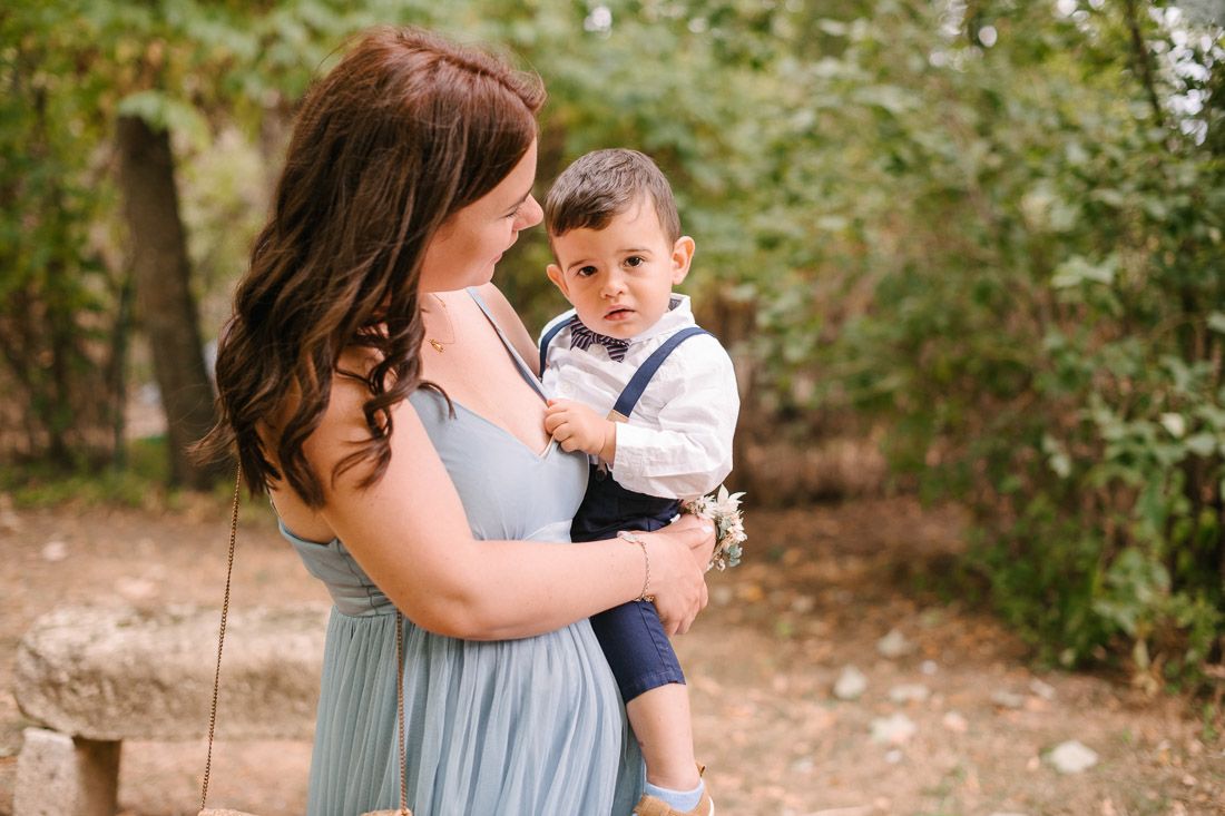 Woman in blue dress tenderly holds a baby boy in suspenders in garden.