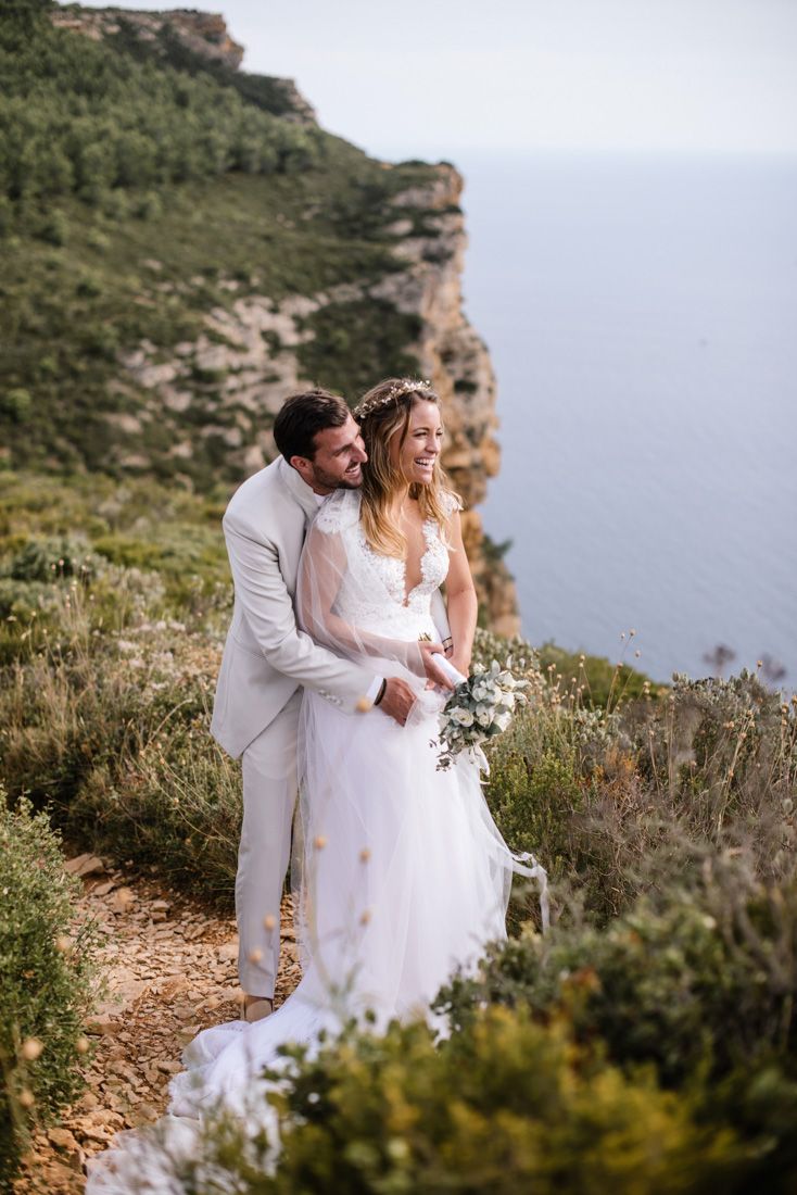 Happy bride and groom embracing on a cliffside path with stunning ocean and rocky cliffs in the background.
