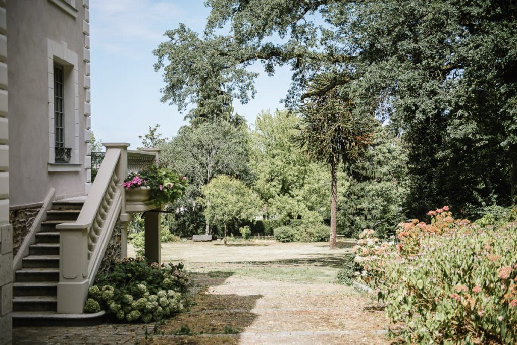 Elegant French chateau garden with lush green trees, manicured grounds, and stone staircase with flower boxes.
