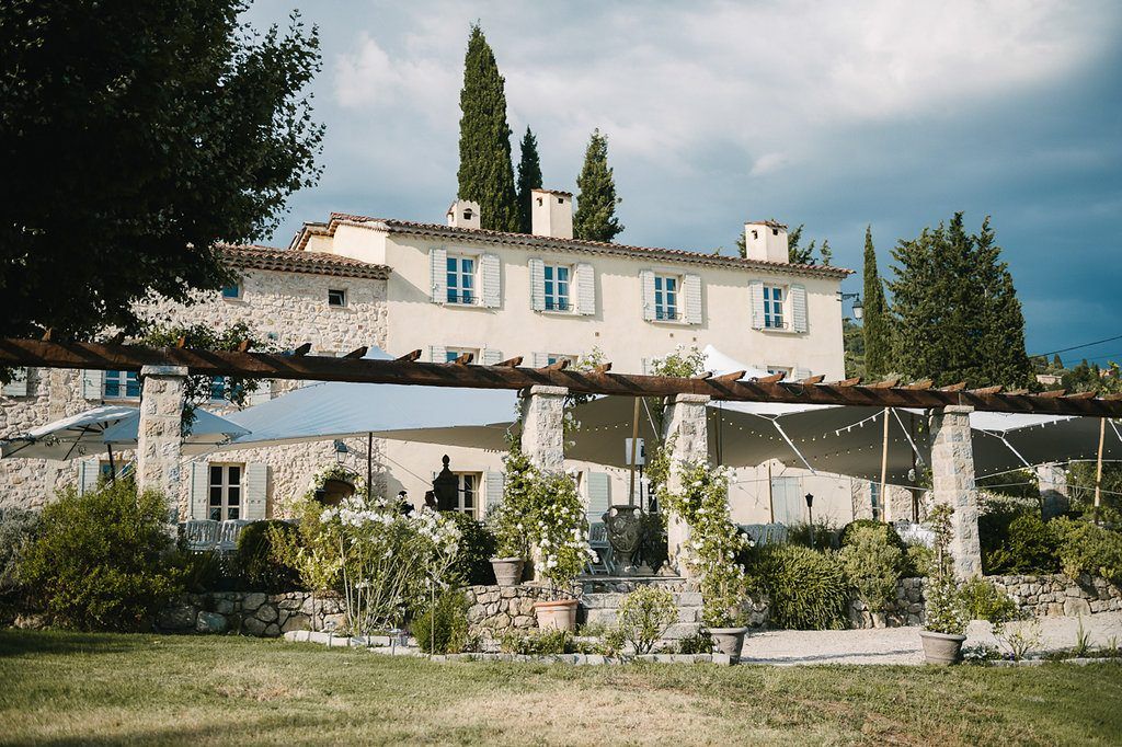 Elegant Provençal stone villa with white shutters, pergola, and lush gardens under a dramatic cloudy sky.