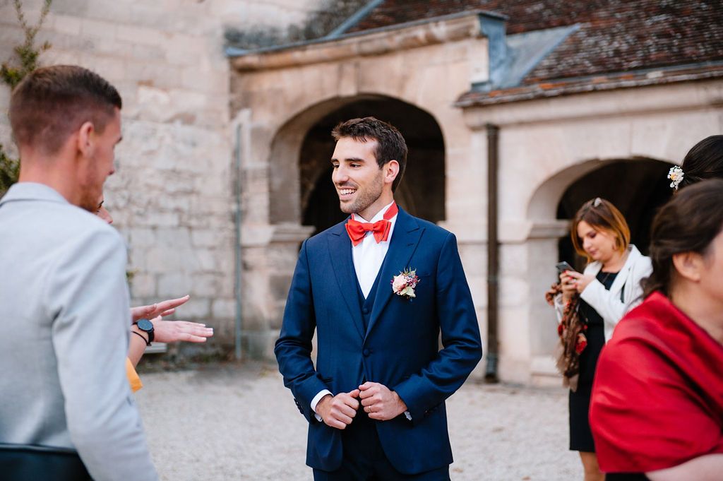 Smiling groom in blue suit with red bow tie greeting wedding guests in a courtyard.