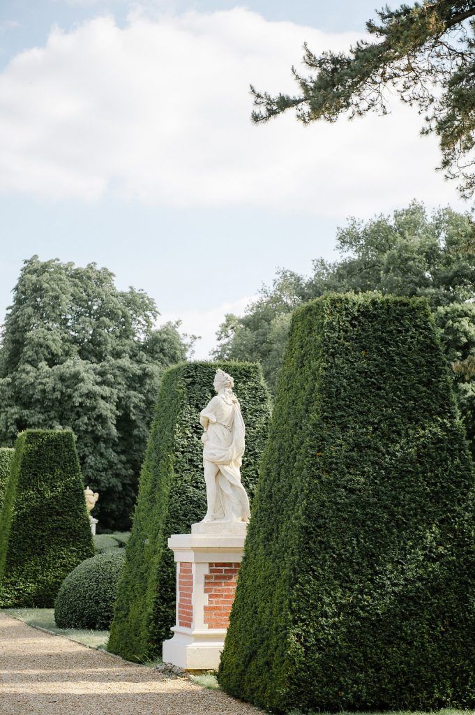 Classical white statue on brick pedestal surrounded by manicured hedges in formal garden.