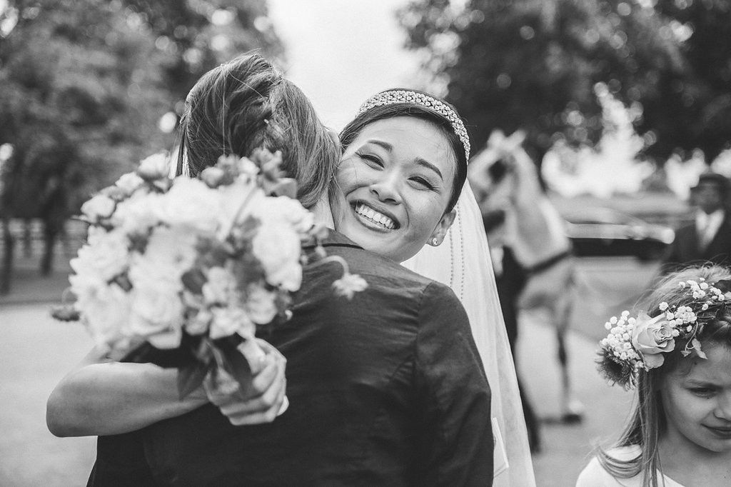 Glowing bride in tiara hugs guest warmly while holding white bouquet as flower girl stands nearby after ceremony.