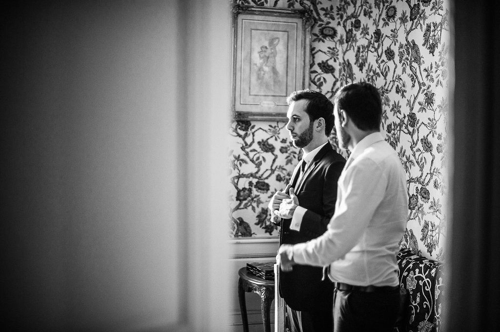 Two men in formal attire conversing in elegant room with ornate wallpaper before wedding.