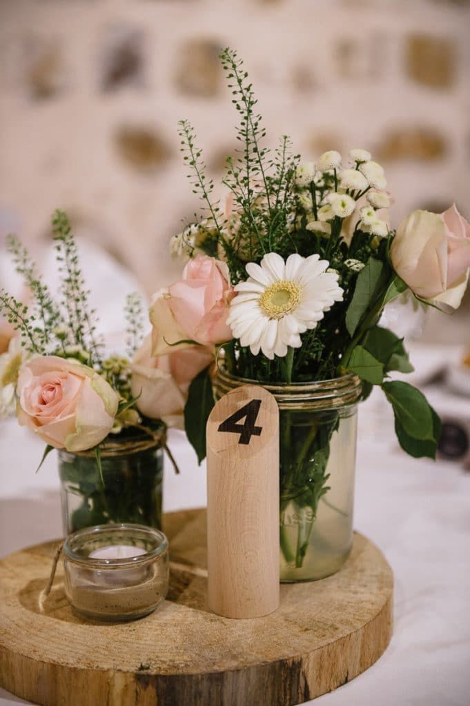 Rustic wedding table centerpiece with pink roses and white gerbera daisies in mason jars on wood.