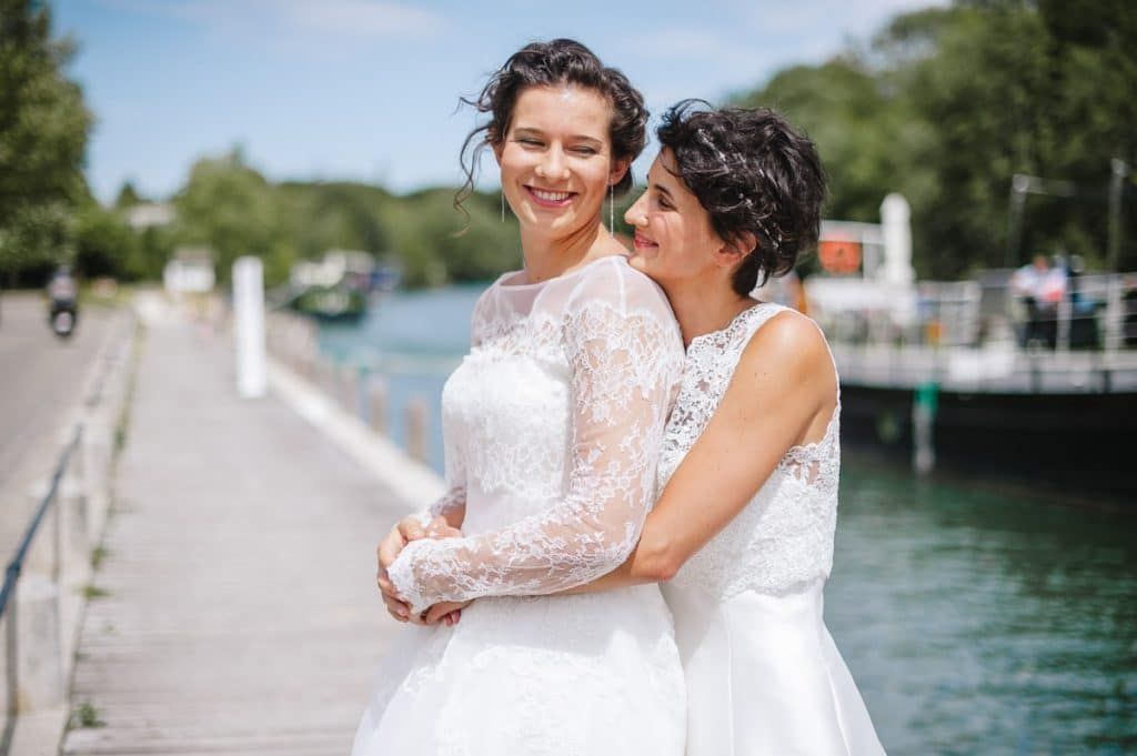 Two brides in white lace wedding dresses embracing near a waterfront.