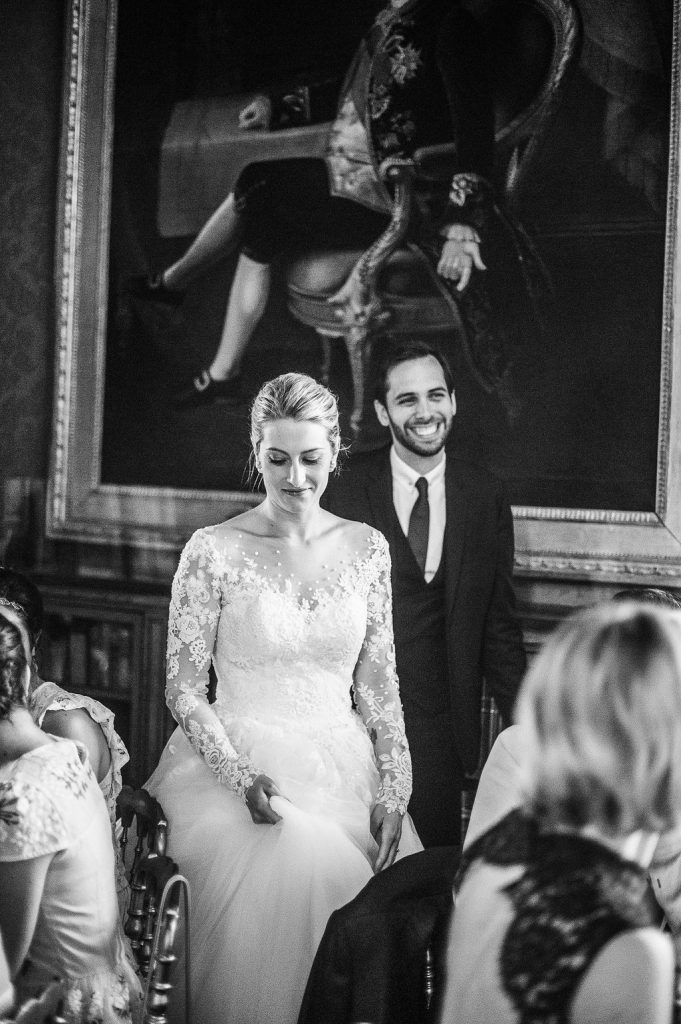 Black and white photo of smiling groom beside emotional bride in lace dress during wedding ceremony.