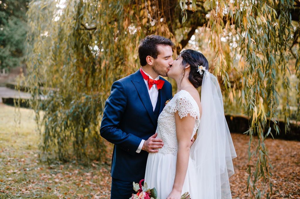 Bride and groom share romantic kiss beneath golden weeping willow branches in fall garden.