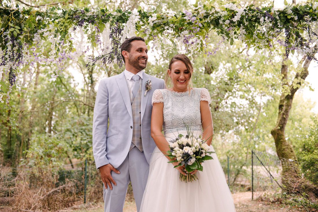 Bride and groom smiling under floral arch in woodland setting, bride holding white bouquet in lace dress.