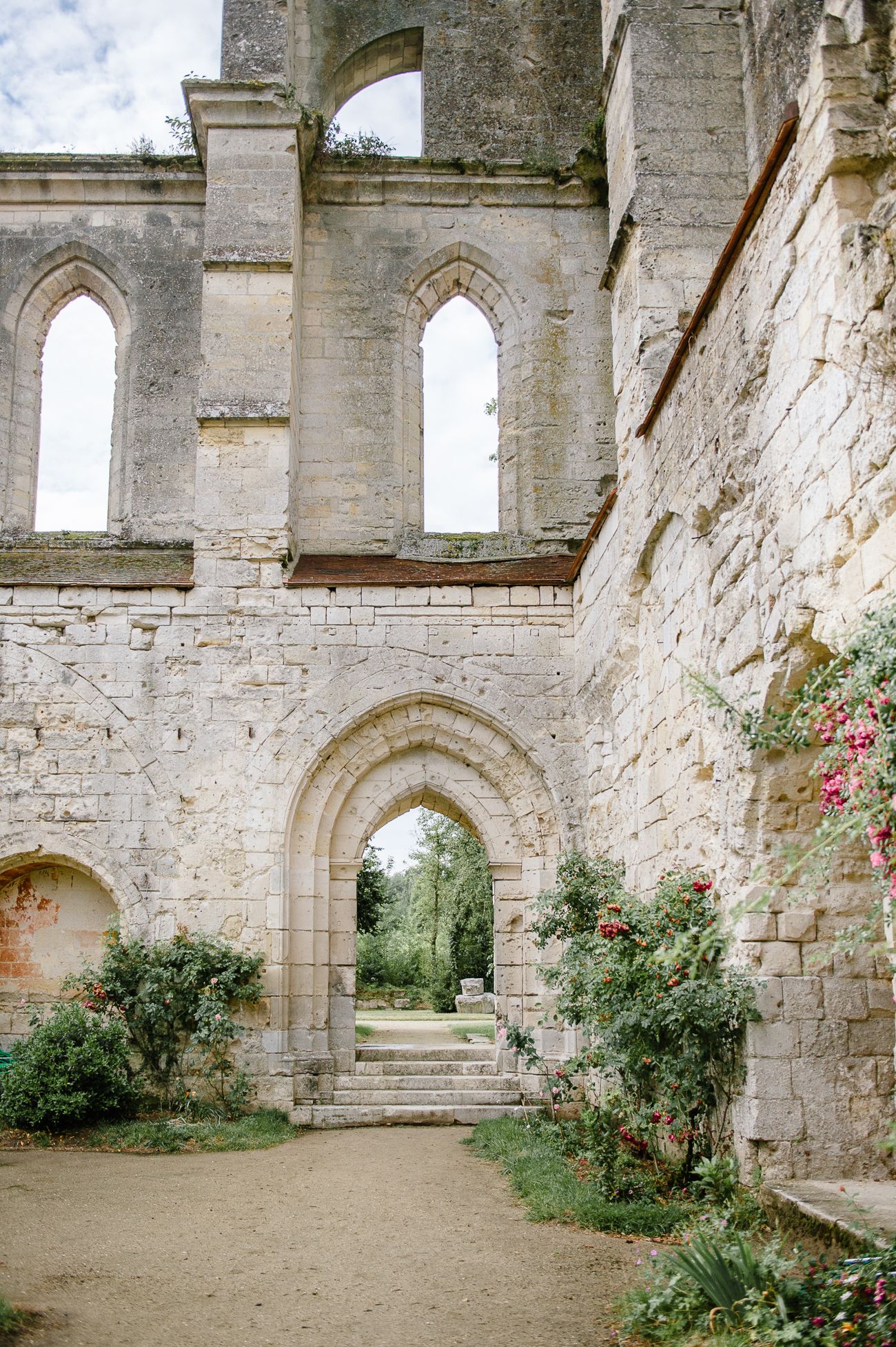 Ancient stone abbey ruins with Gothic arched doorway, steps, and climbing roses in lush garden setting.