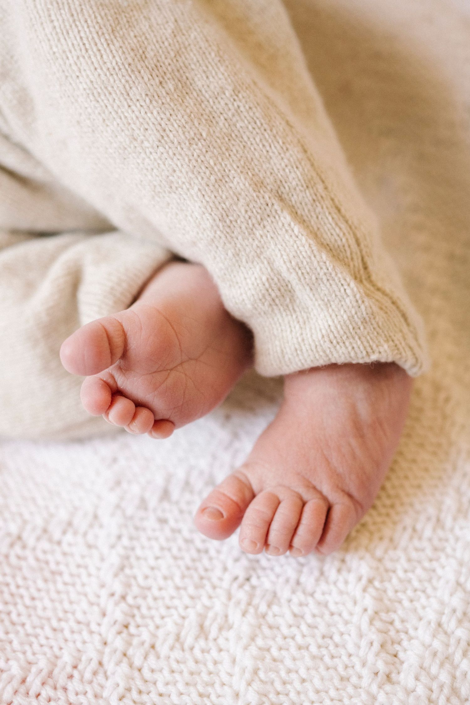 Close-up of tiny newborn baby feet peeking out from a soft beige knit blanket.