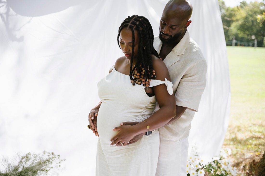 Expecting couple in white outfits embracing outdoors, man tenderly holding pregnant womans belly.