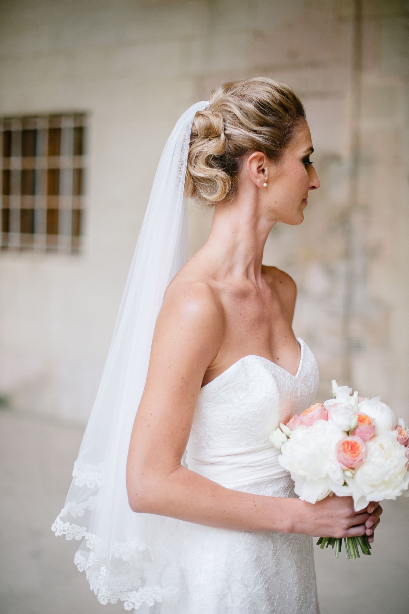 Elegant bride in strapless lace gown holds pink and white bouquet wearing a lace-trimmed veil.