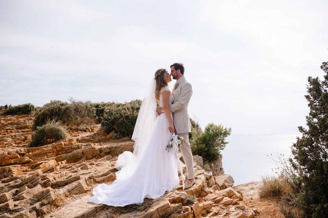 Bride and groom share romantic moment on rocky coastal cliff with ocean backdrop.
