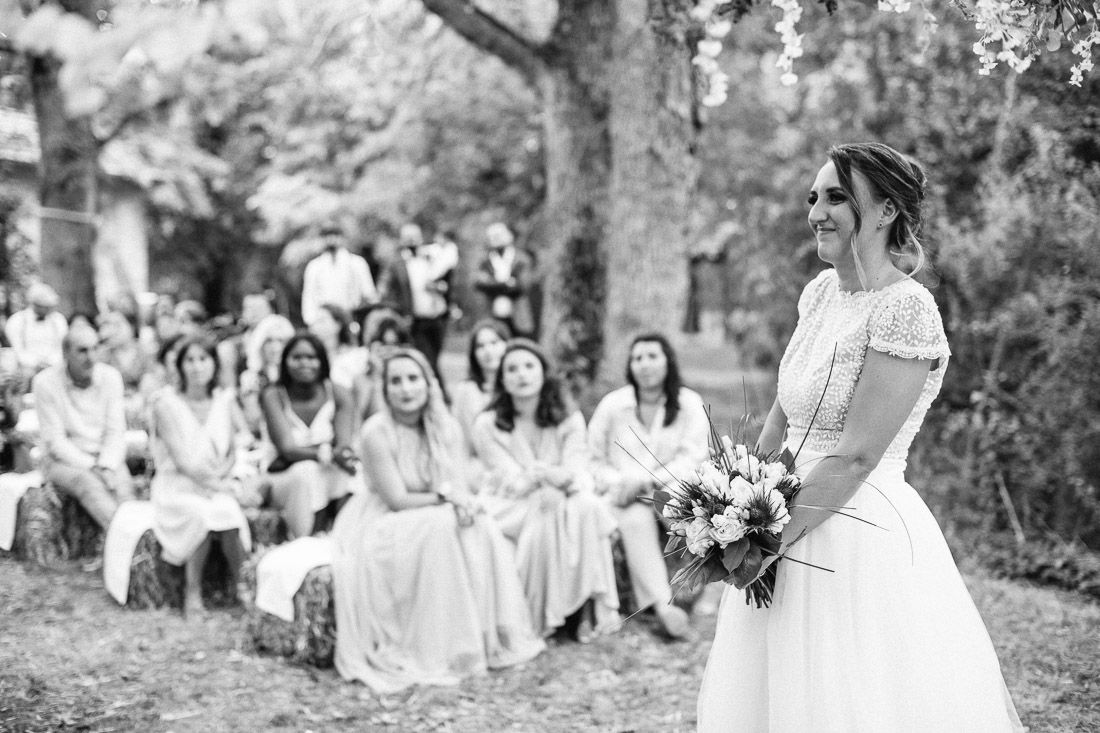 Smiling bride holding bouquet at outdoor wedding ceremony with seated guests in black and white photo.