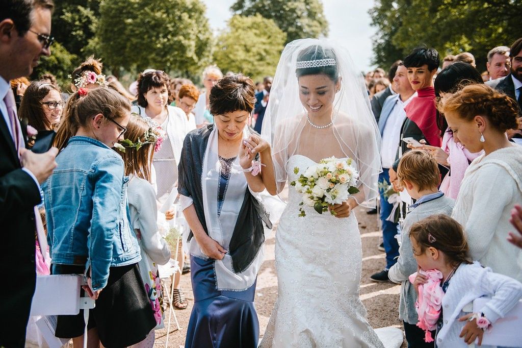 Bride in white lace gown and veil walks with older woman through crowd of wedding guests outdoors.