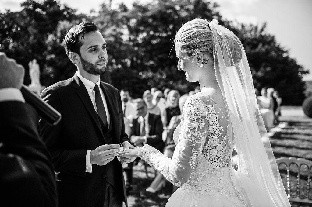 Groom placing wedding ring on bride's finger during outdoor ceremony in black and white photo.