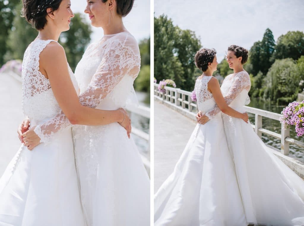 Two brides in lace wedding gowns embrace on a bridge in a romantic outdoor portrait.