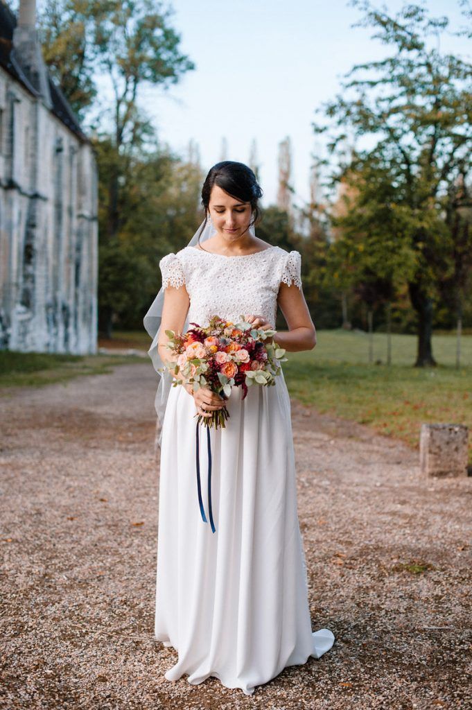 Bride in white lace gown holds vibrant floral bouquet on gravel path near historic estate.