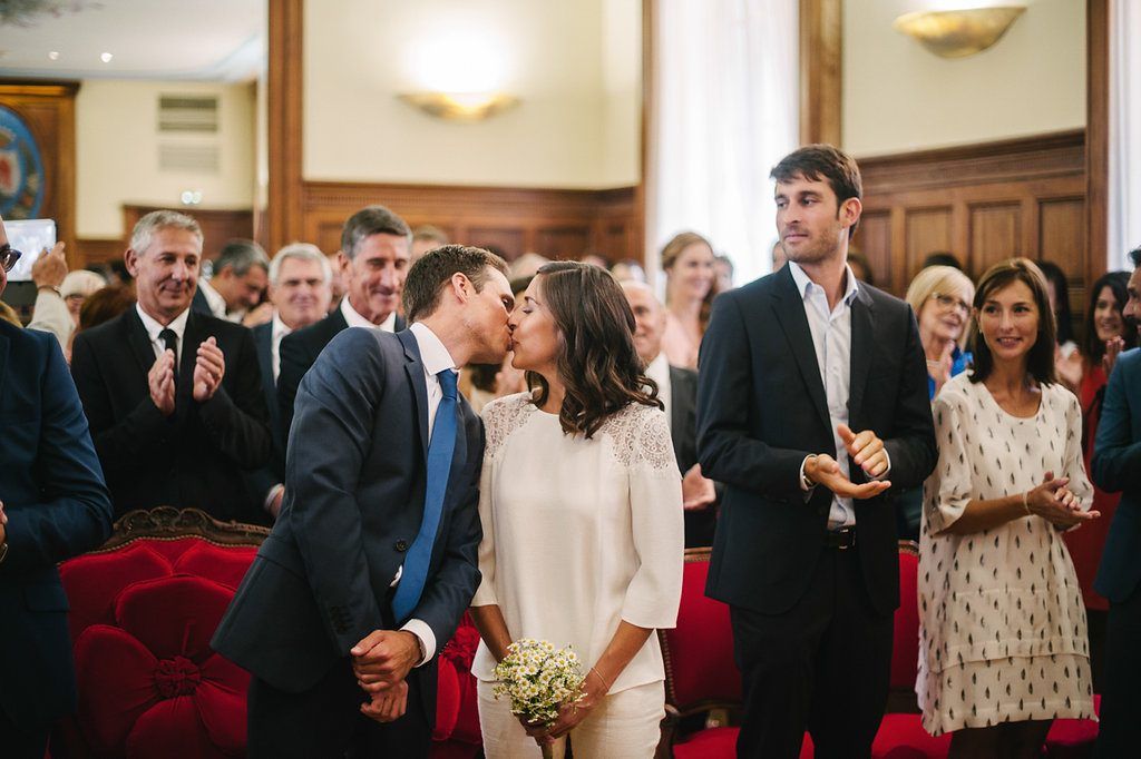 Newlyweds share first kiss at civil ceremony while guests applaud in ornate room.