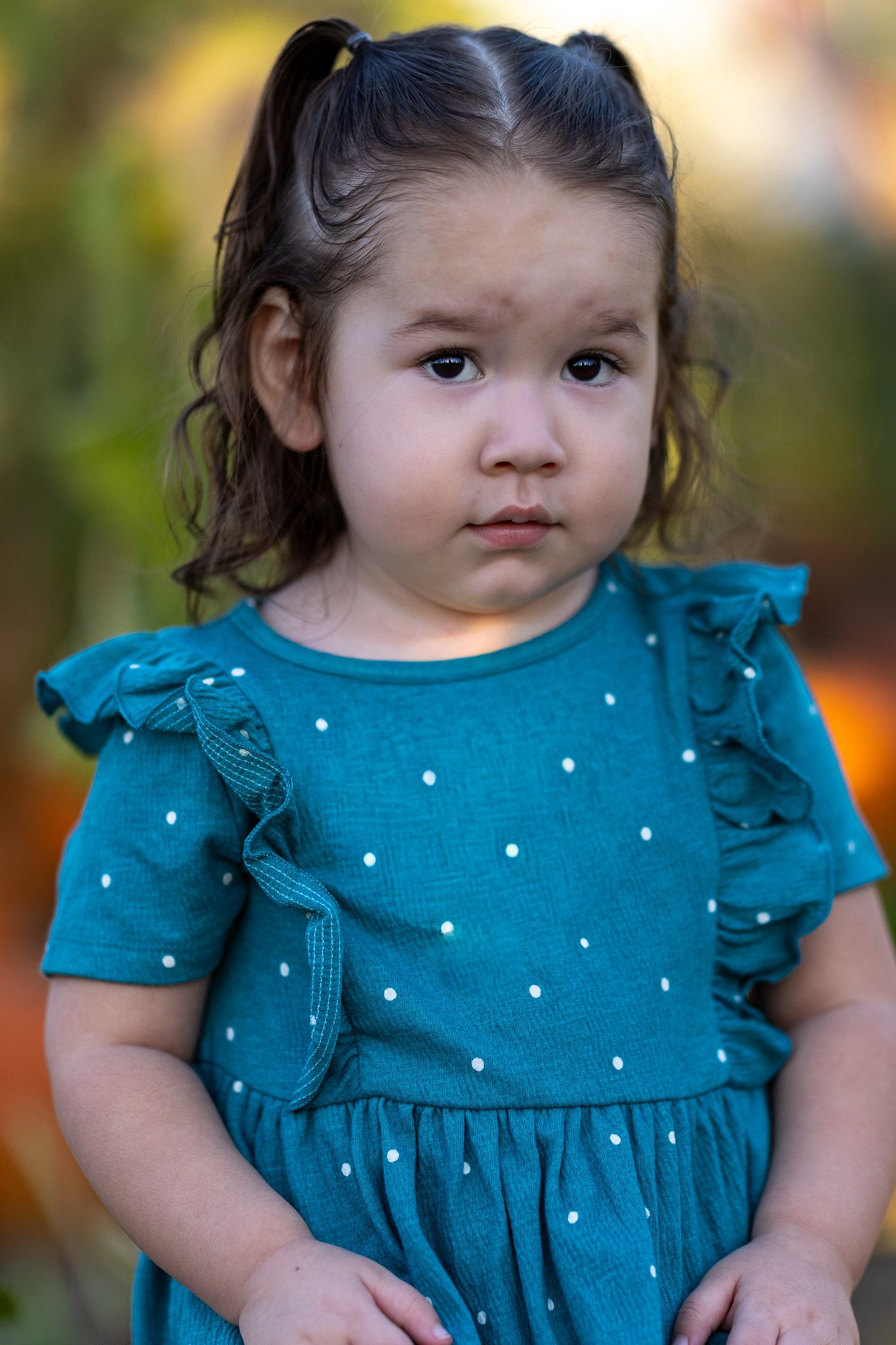 A toddler girl in a teal polka dot dress with ruffle sleeves poses outdoors with a serious expression.