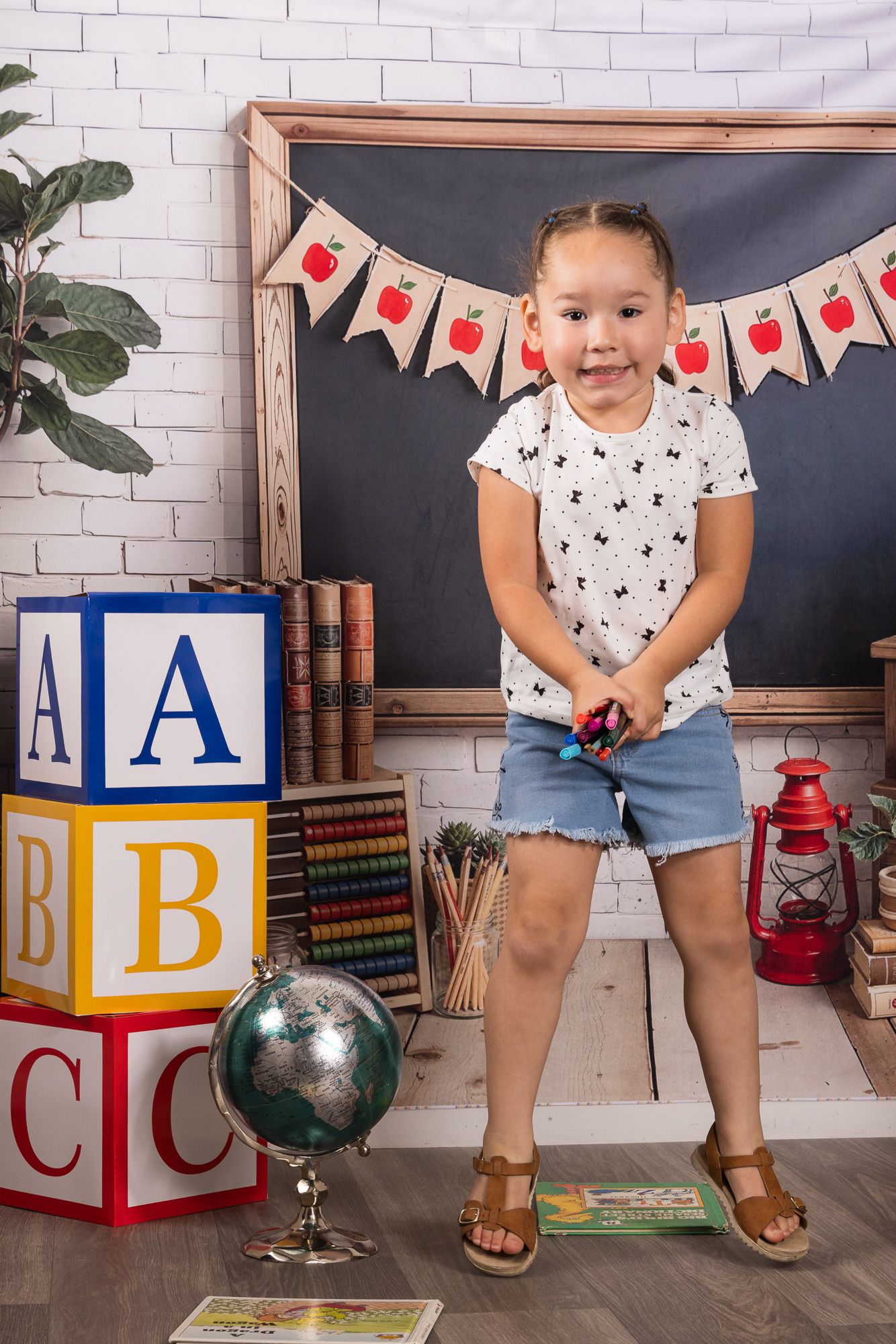 Young girl smiling in a back-to-school photo setup with ABC blocks, globe, books, and chalkboard backdrop.