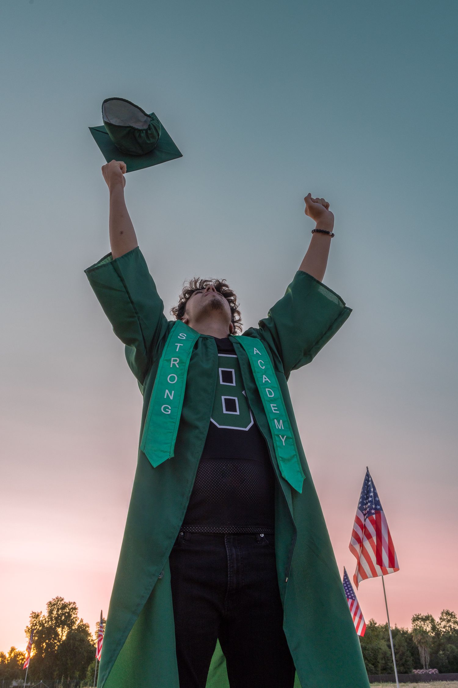 Graduate in green cap and gown raises arms triumphantly at sunset near American flag at Strong Academy.