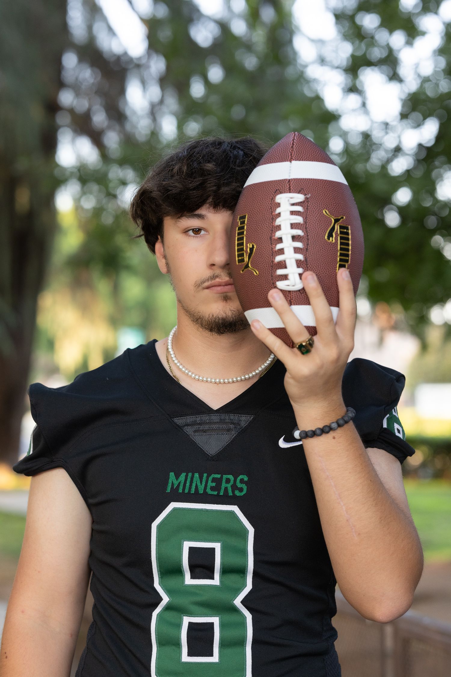 Young football player in black Miners jersey number 8 holds a football up near his face outdoors.