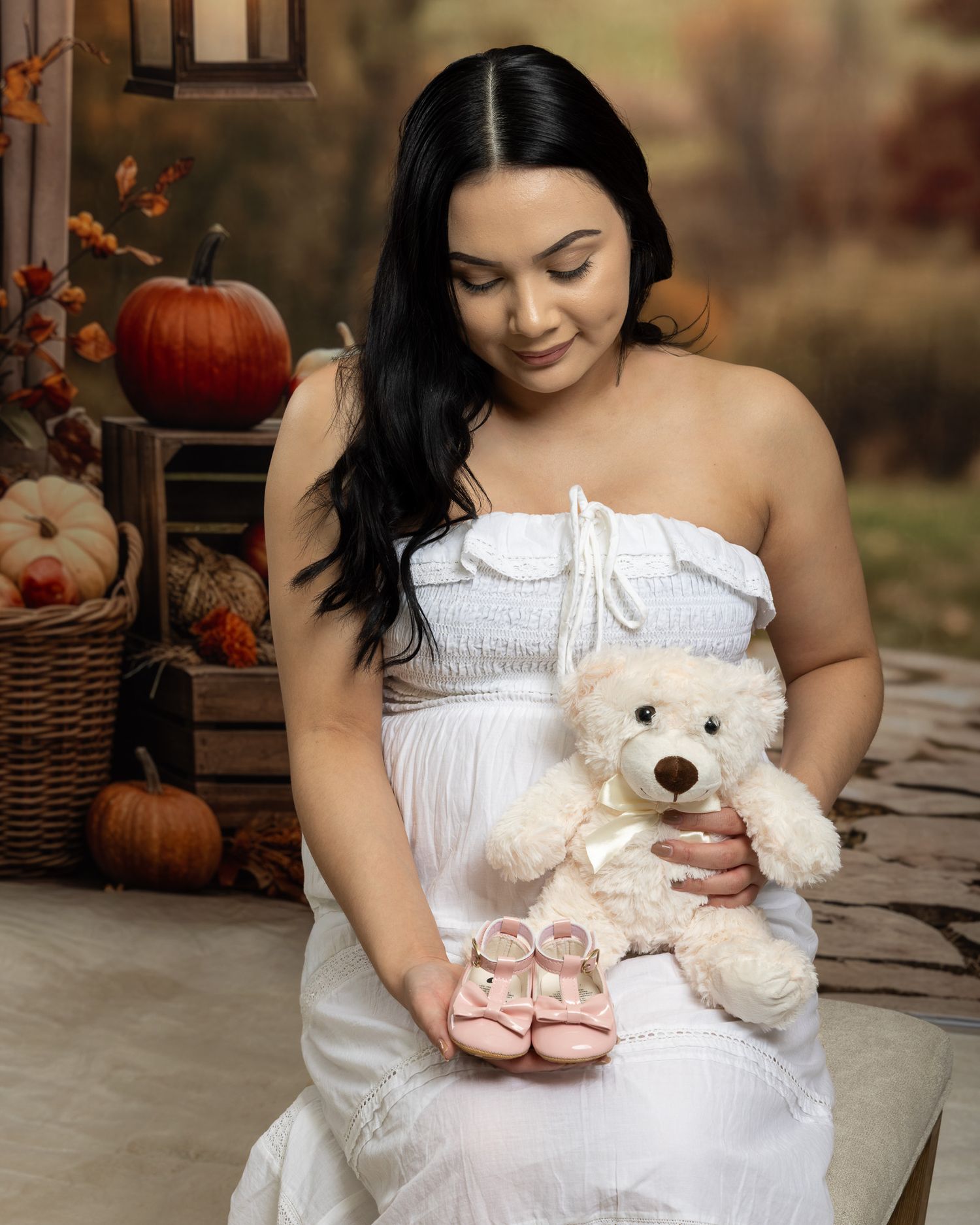 Pregnant woman in white dress holding teddy bear and pink baby shoes in autumn studio maternity photo session.