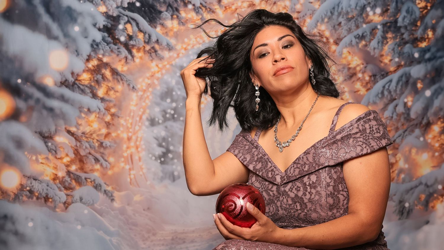 Woman in elegant lace dress holding red Christmas ornament posed before snowy winter backdrop with glowing lights.