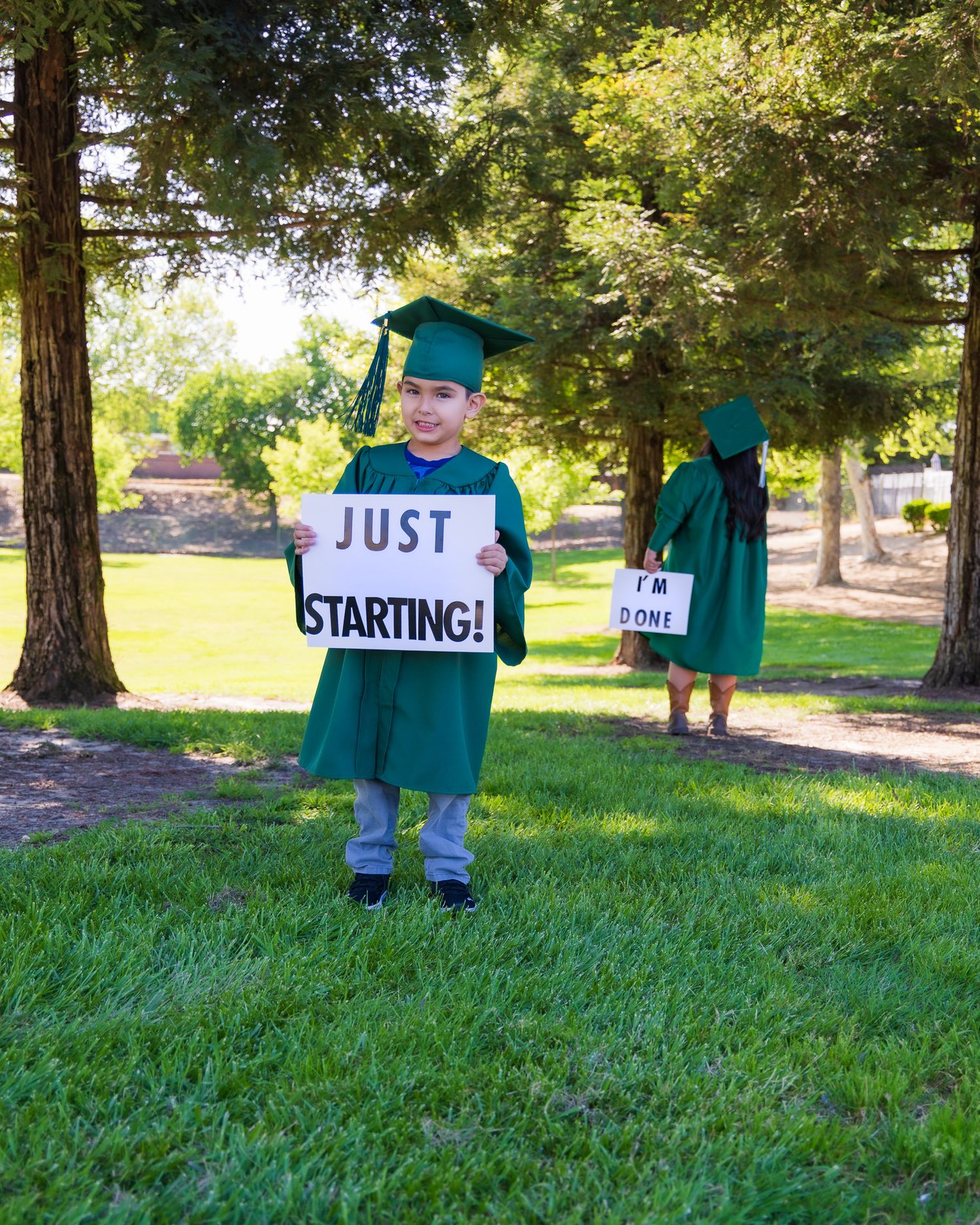 Two children in green graduation caps and gowns hold signs reading Just Starting and I'm Done outdoors.