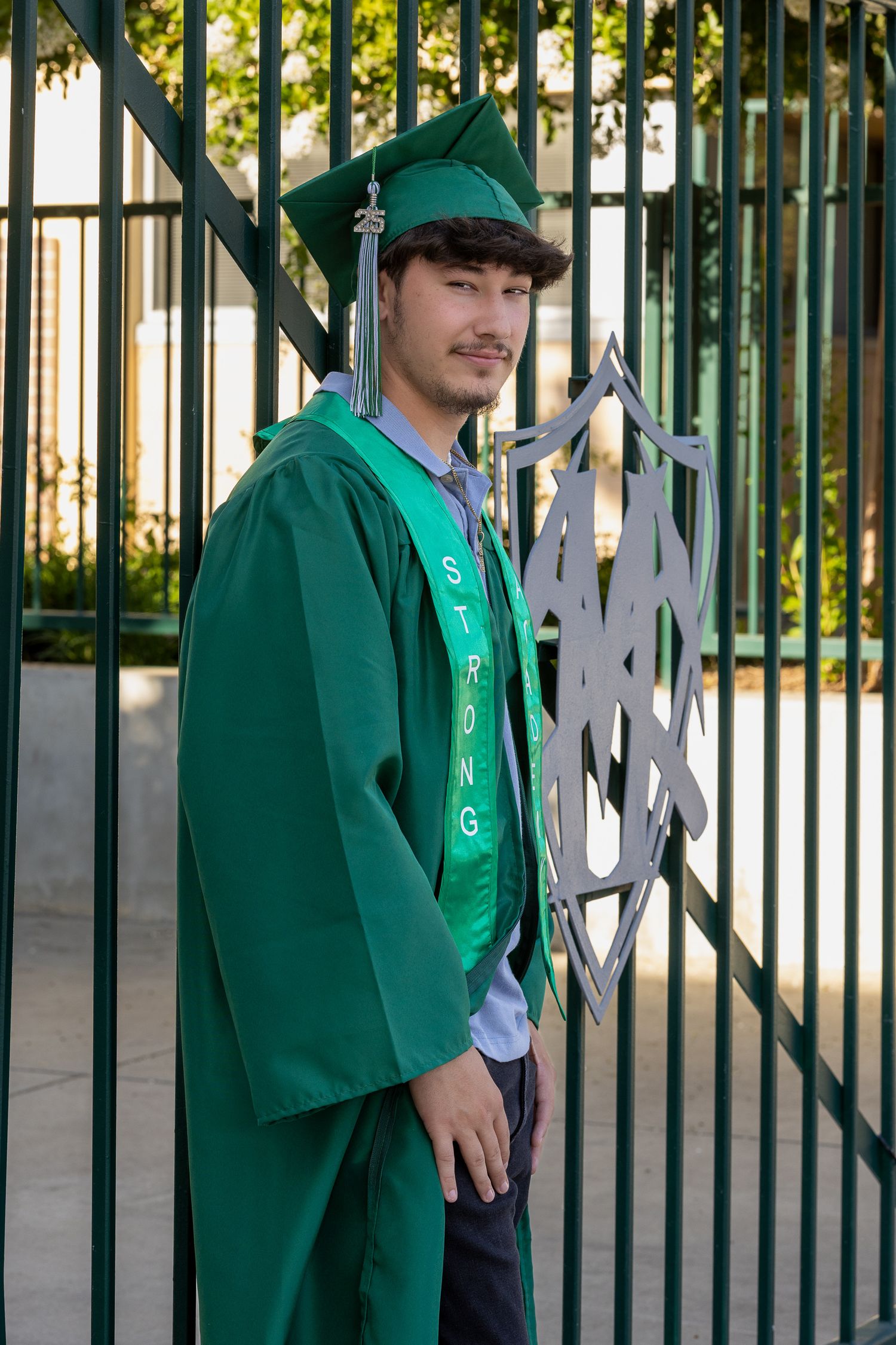 Young man in green graduation cap and gown with STRONG sash poses proudly by decorative metal gate.