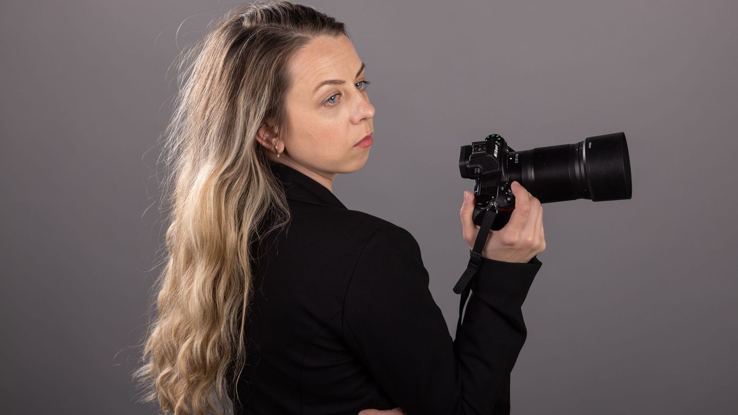 A blonde woman in a black jacket holds a professional DSLR camera with a large lens against a gray background.