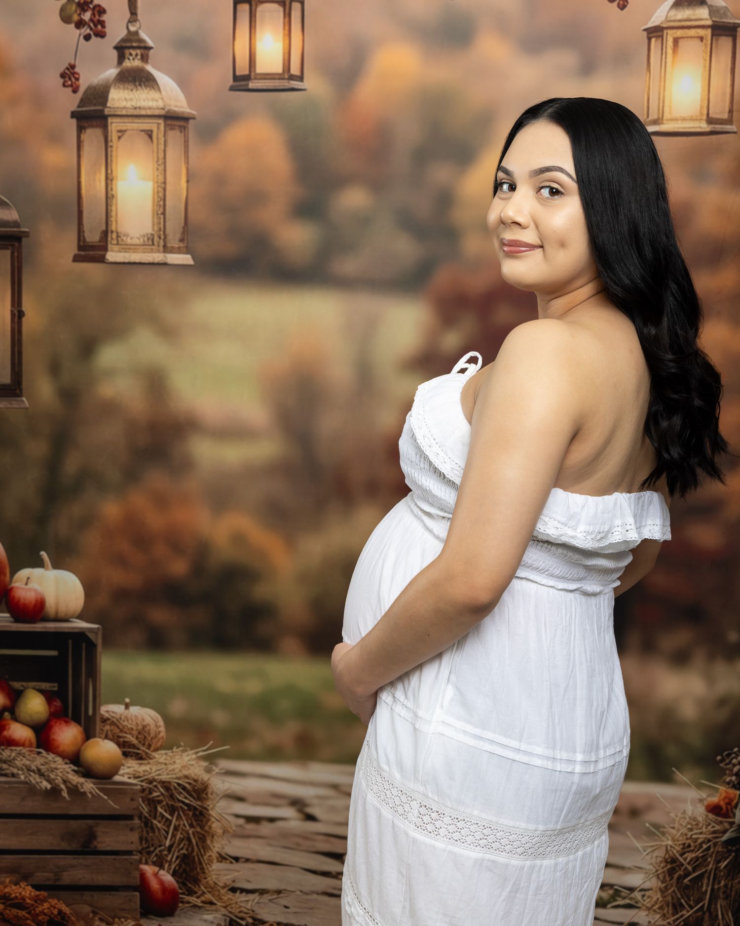 Pregnant woman in white off-shoulder dress poses for fall maternity photo with lanterns and autumn backdrop.