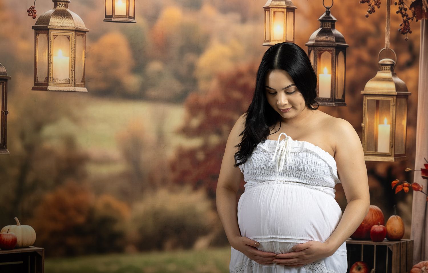 Pregnant woman in white dress poses with autumn backdrop, lanterns, and pumpkins in a maternity photo session.