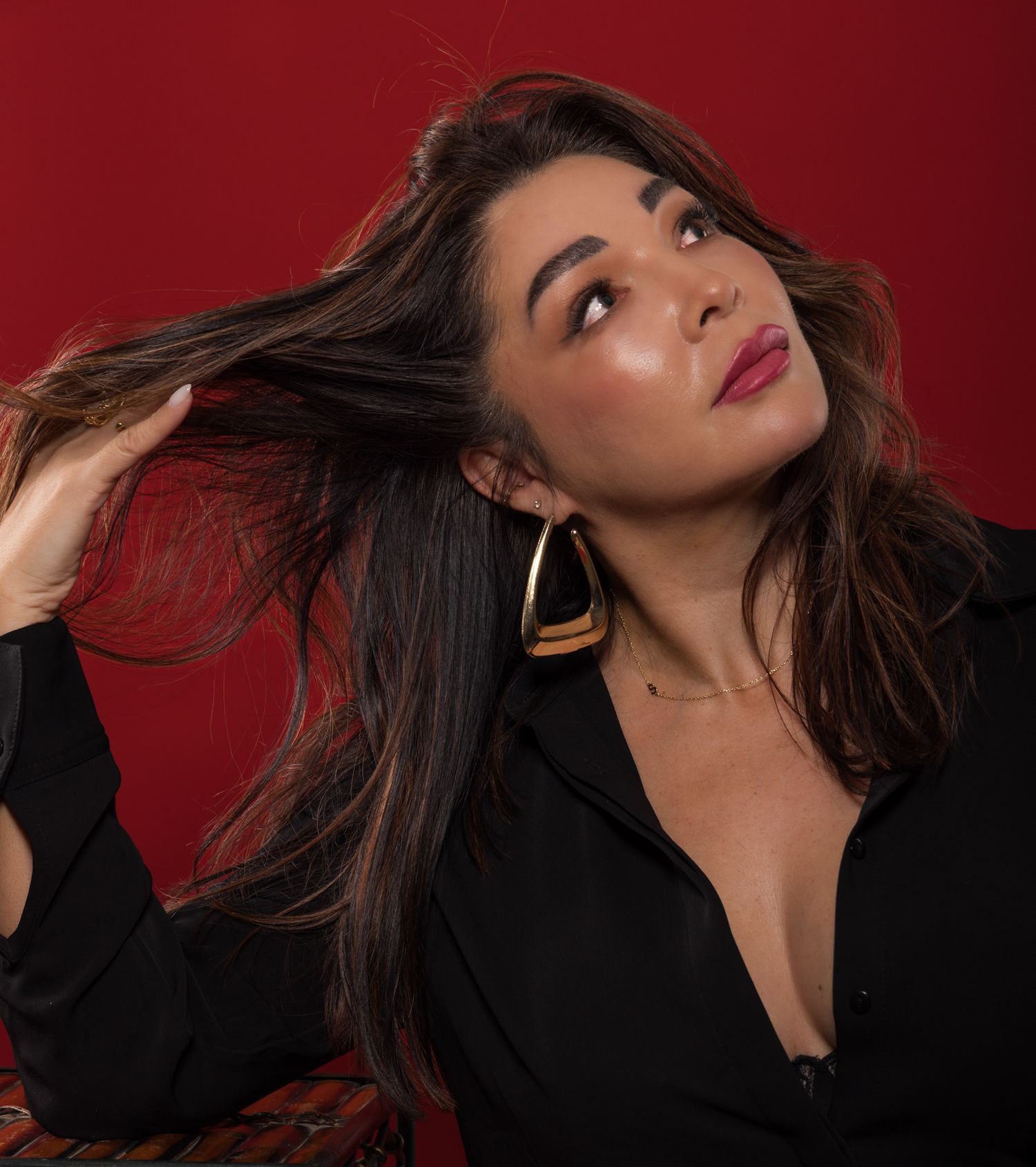 Woman with dark hair in black top poses against red background, wearing gold earrings and bold berry lipstick.