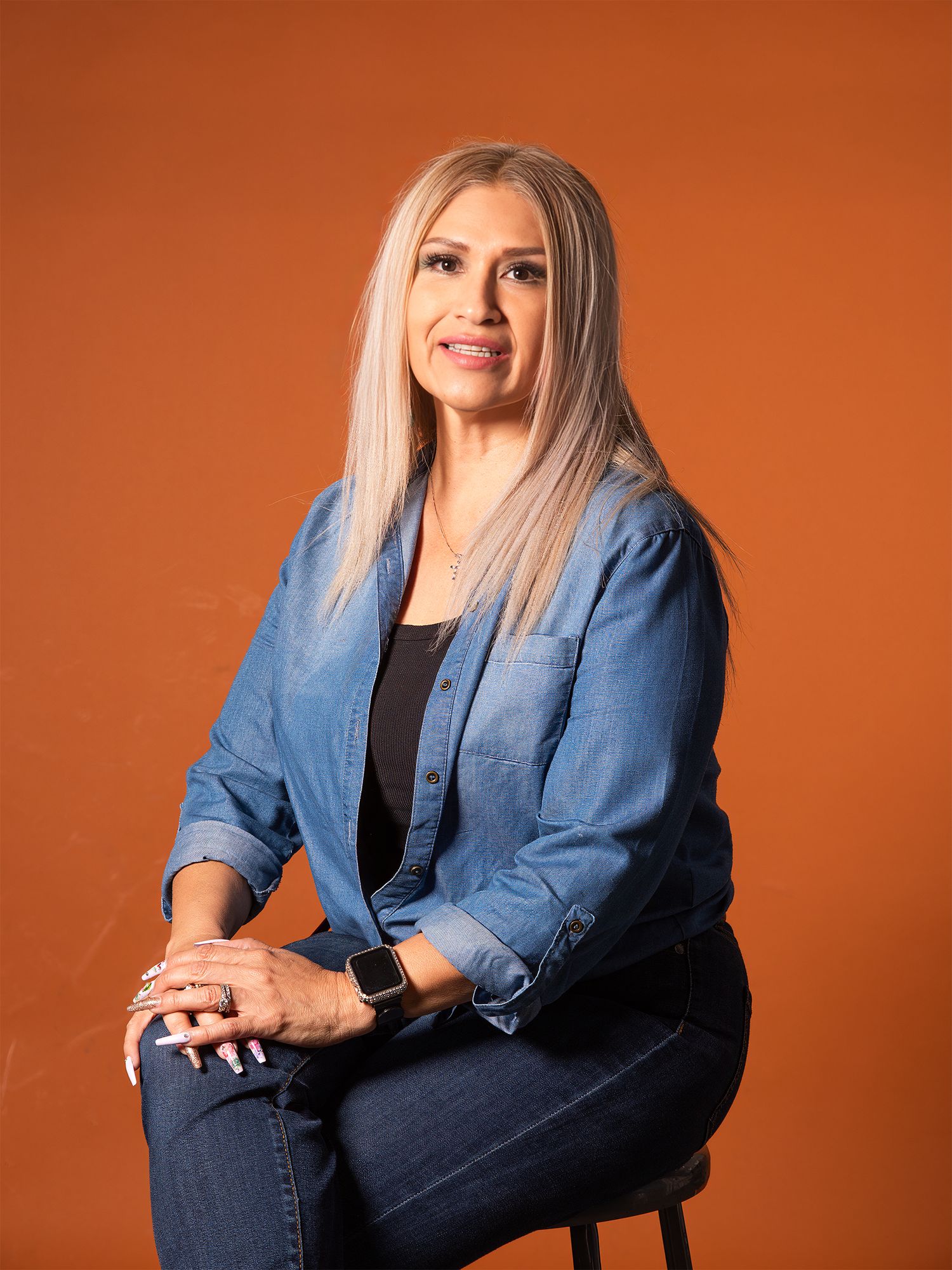 Woman with blonde hair in denim jacket and jeans sits confidently against a warm orange background.