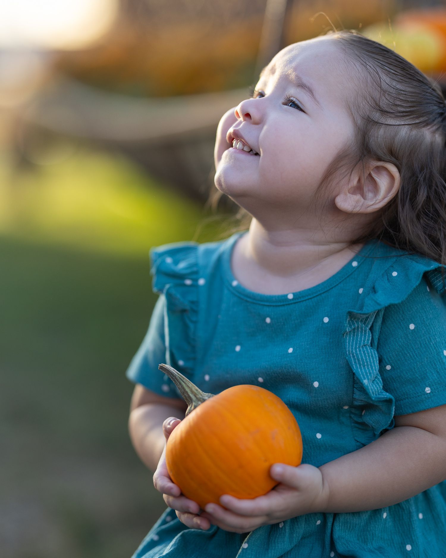 A joyful toddler girl in a teal polka dot dress holds an orange pumpkin outdoors at a fall pumpkin patch.