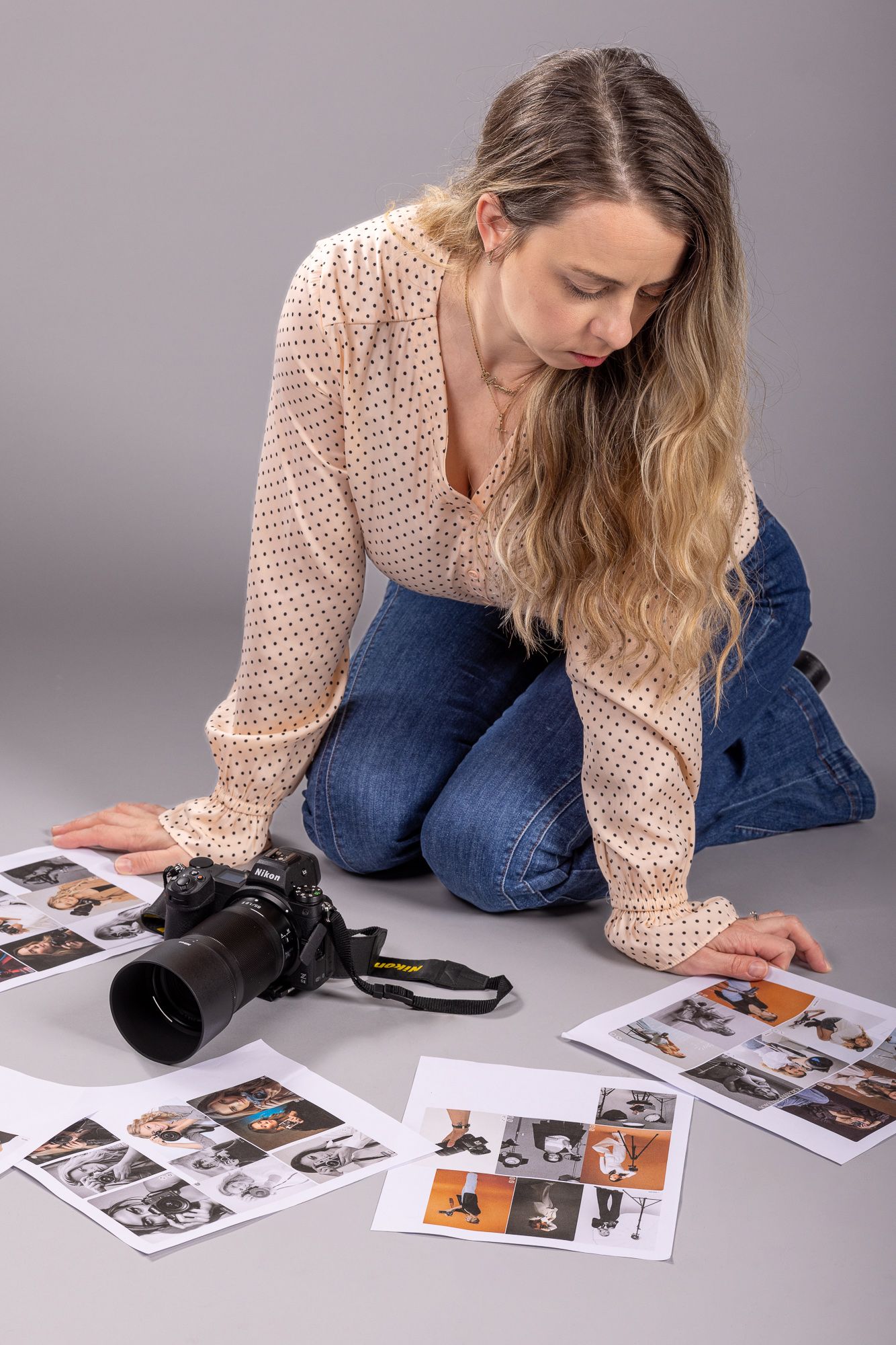 Female photographer kneeling on floor reviewing printed contact sheets beside a DSLR camera on gray background.