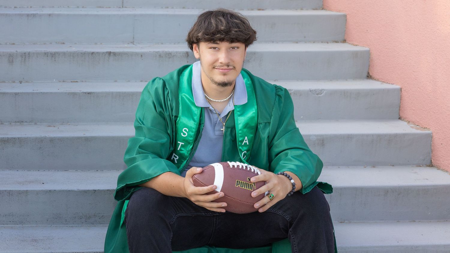 Young man in green graduation gown sits on steps holding a football, celebrating his graduation day.