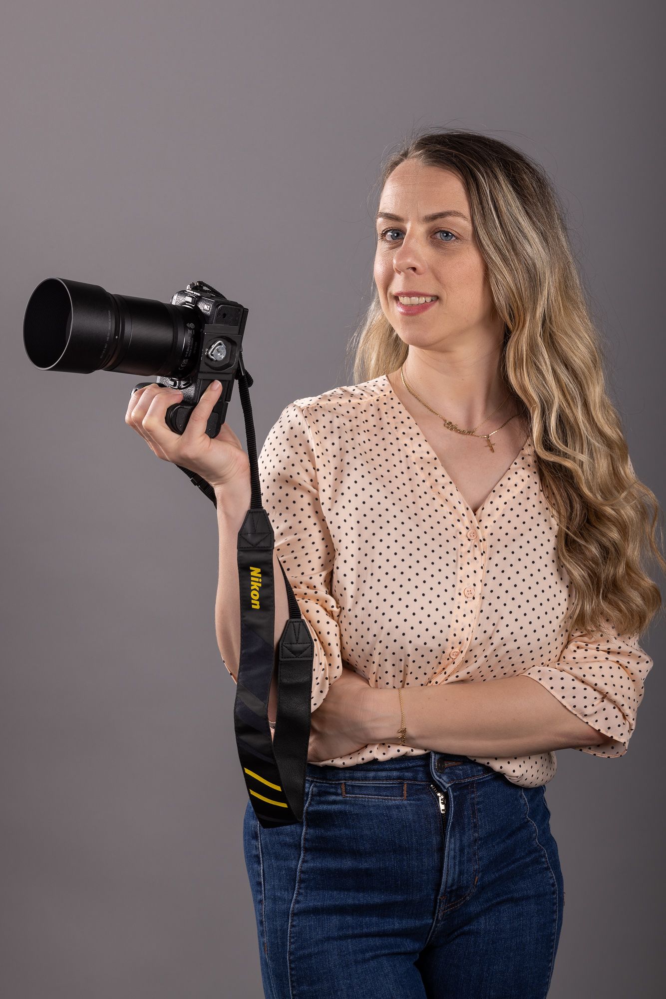Young woman with long blonde hair holding a Nikon DSLR camera with telephoto lens, wearing polka dot blouse and jeans.