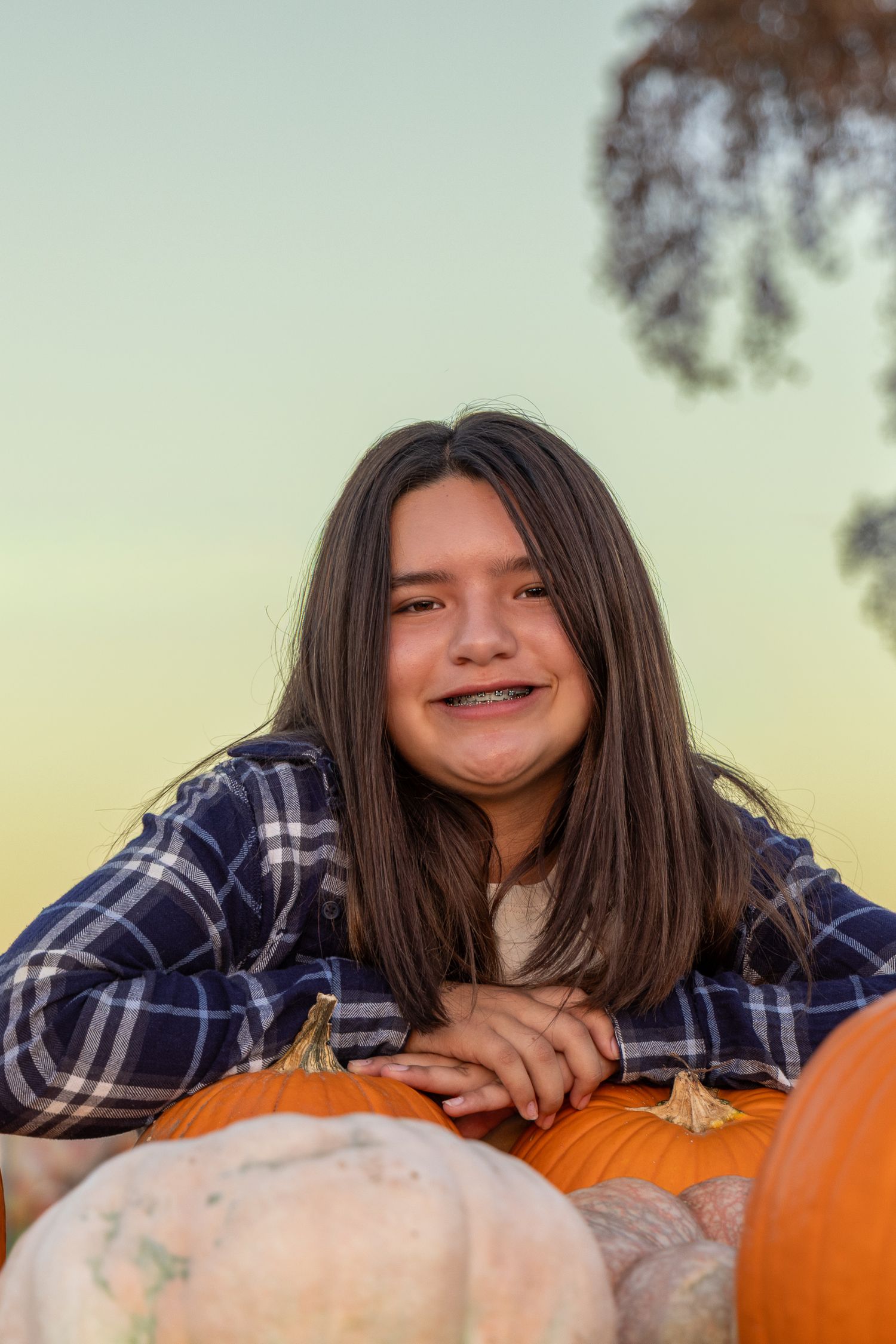 Smiling girl with braces leans on orange pumpkins at a fall pumpkin patch, wearing a blue plaid shirt at sunset.
