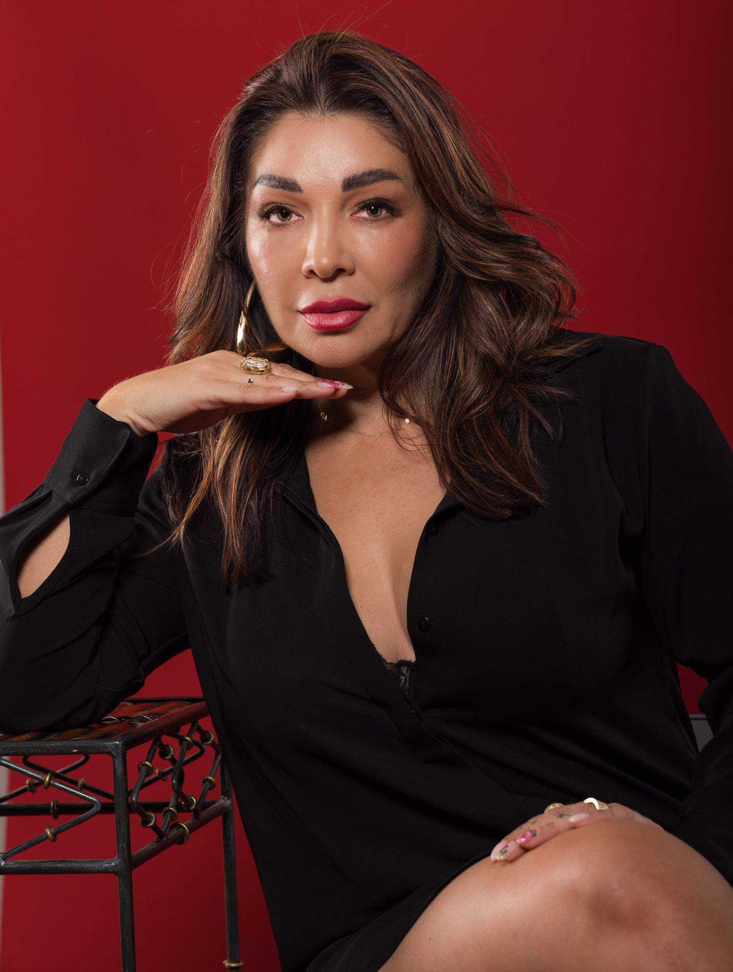 A confident woman in a black dress poses against a red background, showcasing elegant jewelry and flowing brunette hair.
